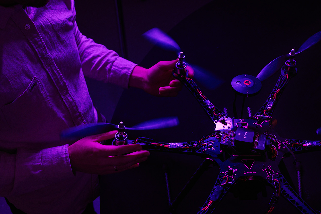 A small multi-limbed robot with propellors and visible circuitry, held in the hands of a person interacting with it.