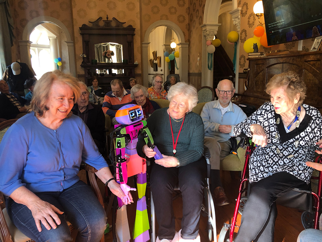 Four elderly people in an aged care facility gathered around a brightly-coloured humanoid robot.