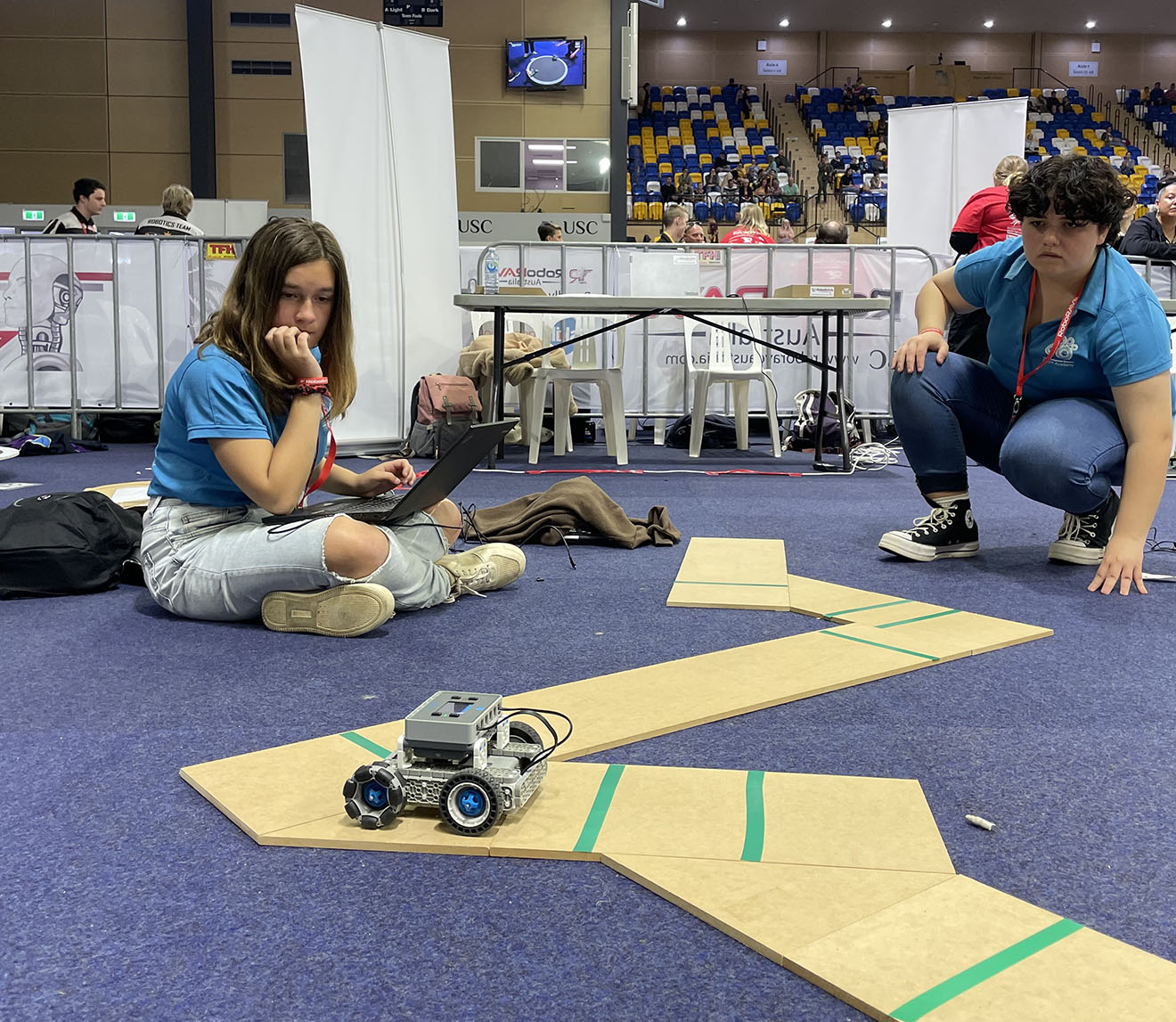 Two young people in a carpeted arena. They are using a laptop to monitor a remote-controlled car-shaped robot on an unusually shaped track.