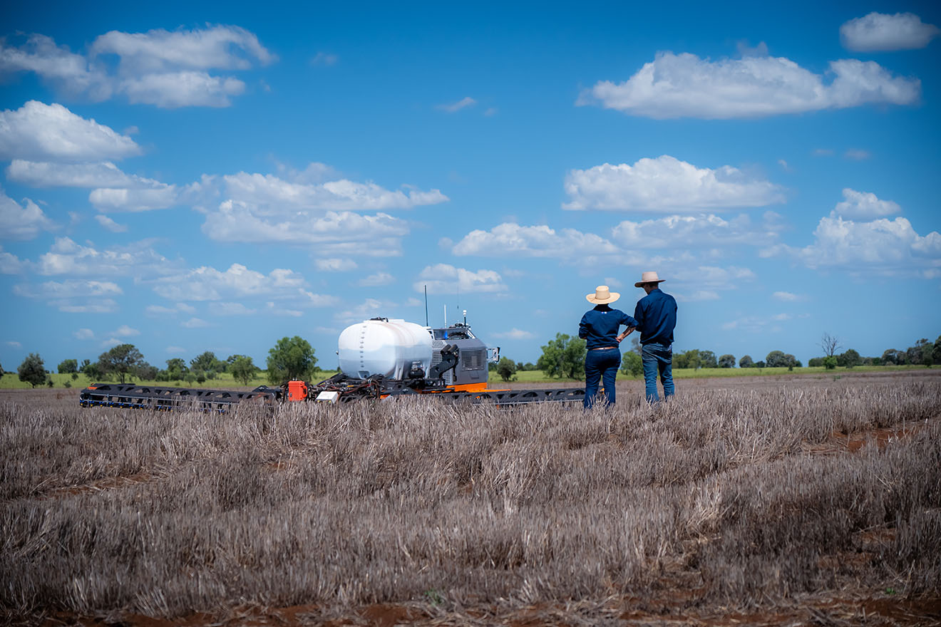 Two farmers standing in a field looking at a large white robot that is working autonomously on their crops.