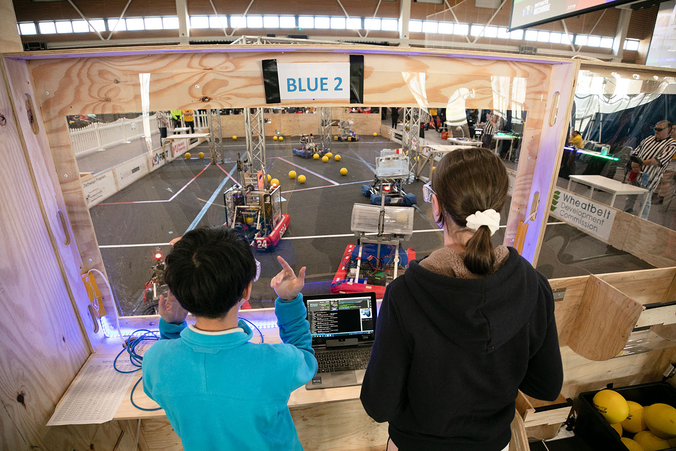 Two young people using remotes to control robots in an arena.