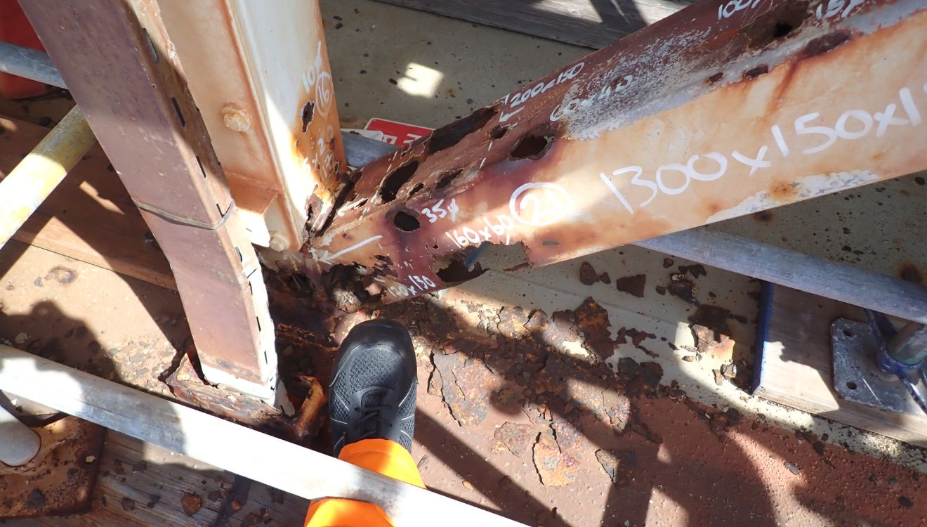 Photo of a worker's boot pointing to heavily rusted support beams.