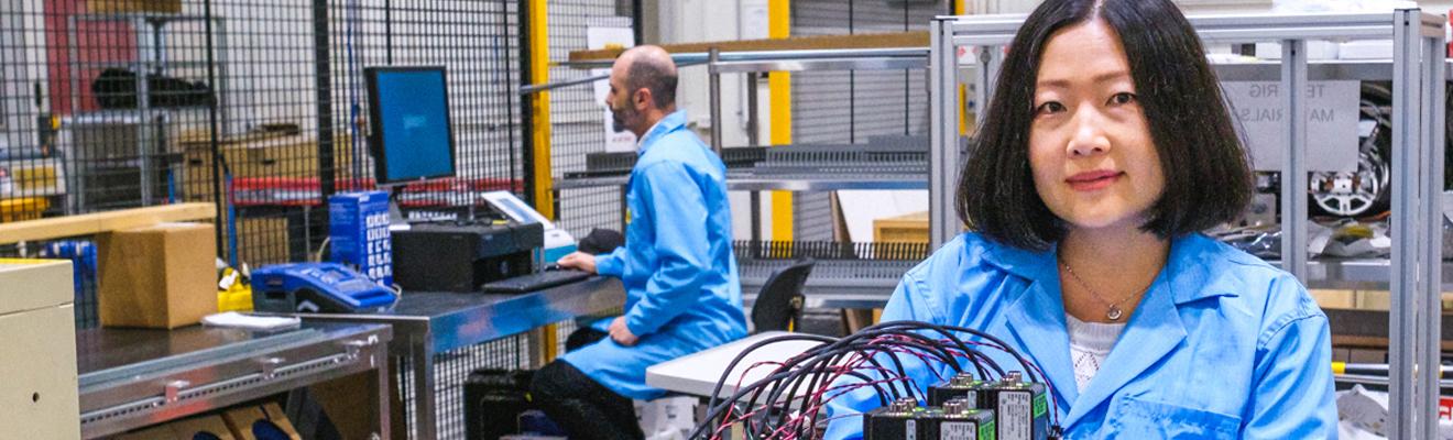 Two researchers in a lab. In the background a man is sitting at a computer and in the foreground a woman is looking at the camera while working on a battery device.