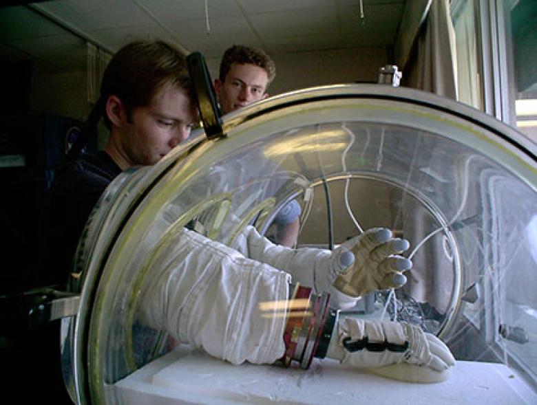 Two technicians work with their arms within a hypobaric chamber which is being used for testing Skinsuit gloves for spacewalking.