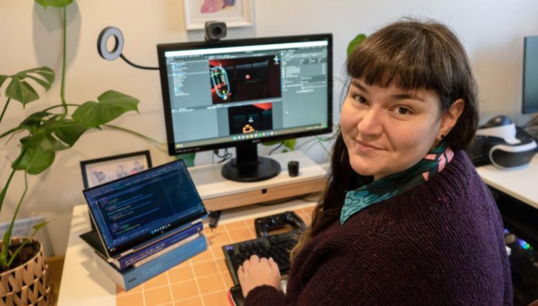 Photo of Cherie at a desk with 2 screens, a keyboard and a games controller. One screen is showing computer code, the other 3D modelling software