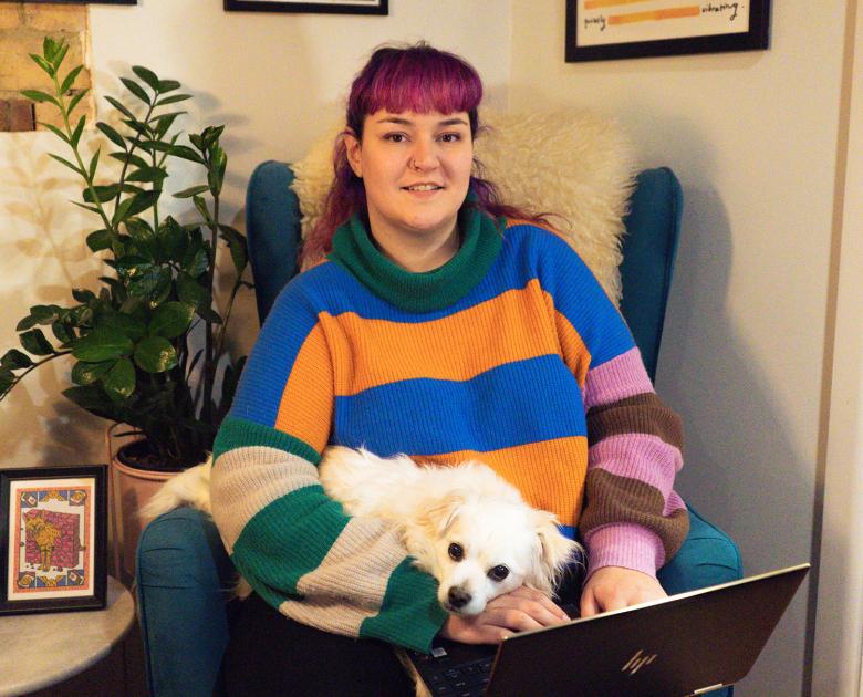 Photo of Cherie using a laptop while sitting on an armchair. A small white dog is on her lap.