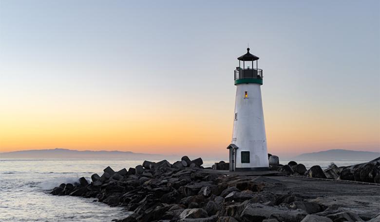 Photo of a lighthouse on a rocky outcrop by the sea at sunset.