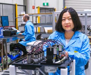 Two researchers in a lab. In the background a man is sitting at a computer and in the foreground a woman is looking at the camera while working on a battery device.