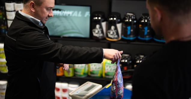 Photo of an NMI staff member weighing a package on electronic scales. A person stands in front of the scales to the right of the screen.