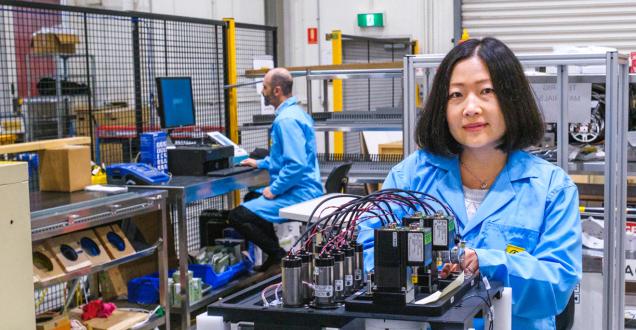 Two researchers in a lab. In the background a man is sitting at a computer and in the foreground a woman is looking at the camera while working on a battery device.