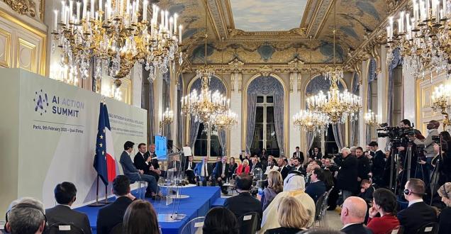 Photo of the AI Action Summit stage located in the Grand Palais. Several political figures are sitting down and talking to a room filled with people. 