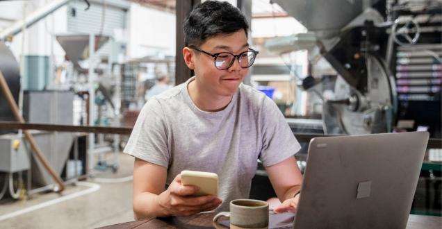 A young man on his laptop with a phone handy. He's sitting in a tech lab with an office behind him.