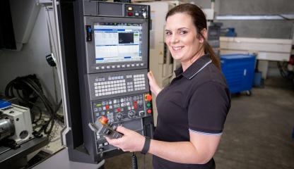 Image shows a woman in a factory. She is standing next to the control panel for a machine tool, smiling and looking towards the camera.