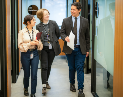 Three staff from diverse backgrounds walk down a corridor looking at each other smiling. 