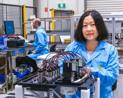 Two researchers in a lab. In the background a man is sitting at a computer and in the foreground a woman is looking at the camera while working on a battery device.